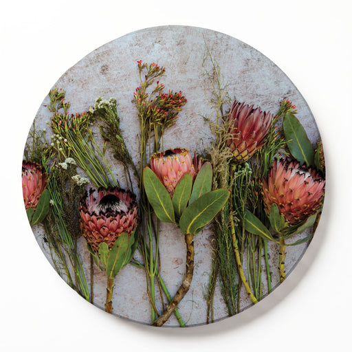 A round wooden serving board featuring a botanical print of pink and red protea flowers and green foliage on a textured light grey background, shown against a clean white backdrop.