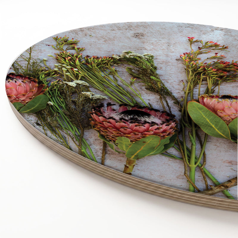 A round wooden serving board featuring a printed botanical design of pink and red protea flowers and green foliage on a weathered light grey background, photographed on a clean white surface.