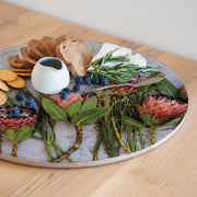 A round serving board with a red protea print styled with bread, cheese, blueberries, crackers, greens, and flowers, set on a light wooden table.