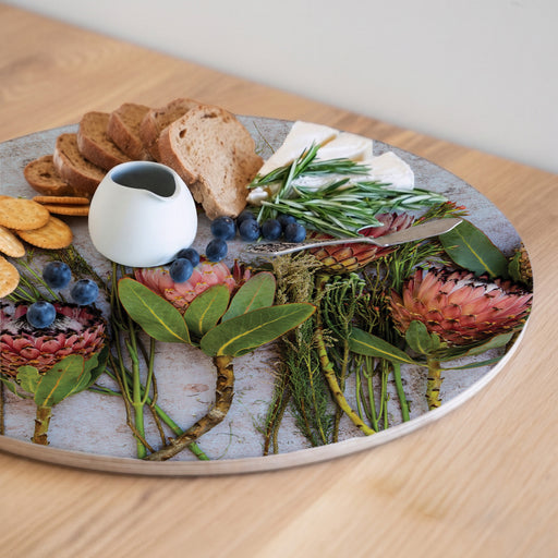 A round serving board with a red protea print styled with bread, cheese, blueberries, crackers, greens, and flowers, set on a light wooden table.
