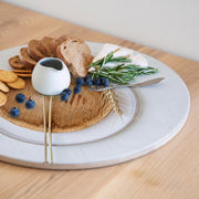A round wooden serving board featuring a beige wheat-on-wood print on a light wood table, featuring a white ceramic pitcher, blue cheese, sliced bread, crackers, blueberries, and rosemary.