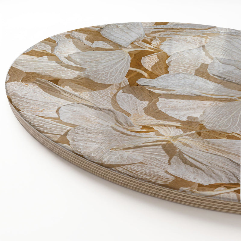 A close-up of a round wooden serving board featuring a bleached cream hydrangea floral pattern in warm tan and white tones, photographed on a clean white background.