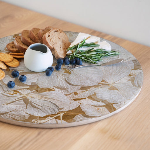 A round serving board with a bleached cream hydrangea floral print pattern, styled with bread, crackers, blueberries, cheese, rosemary, and a small white pitcher on a wooden table.