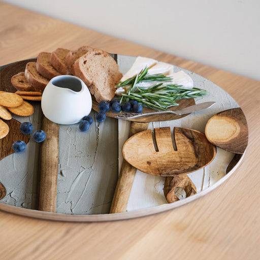 A round wooden serving board featuring a brown wooden spoon print with a charcuterie board arrangement, including bread slices, blueberries, a small white pitcher, cheese, rosemary, crackers, set on a light wood tabletop.