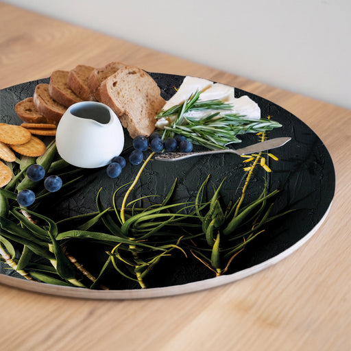 A round wooden serving board featuring a photographic print of aloe branches with yellow blossoms, styled with crackers, bread, blueberries, cheese, green beans, rosemary, and a small white ceramic pitcher on a light wood table.